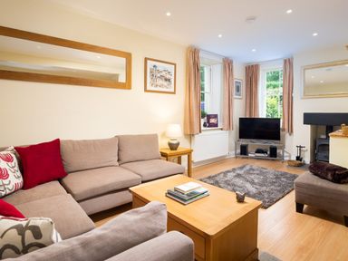 A living room with a beige sectional sofa red cushions a wooden coffee table books a TV on a metal stand and windows with curtains at Scots Pine in Keswick