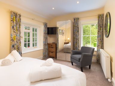 A bedroom with a bed rolled towels a gray armchair mirrored wardrobe two windows with floral curtains and a small wooden drawer unit at Scots Pine in Keswick