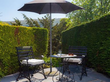 A small outdoor patio with two chairs and a round table under a large umbrella surrounded by tall hedges at Corner Cottage in Troutbeck