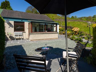 An outdoor patio with chairs a table and umbrella in front of a cottage and garden at Corner Cottage in Troutbeck