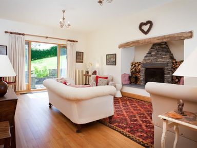 A living room with two white sofas a stone fireplace and a glass door showing a garden at The Old Coach House in Troutbeck
