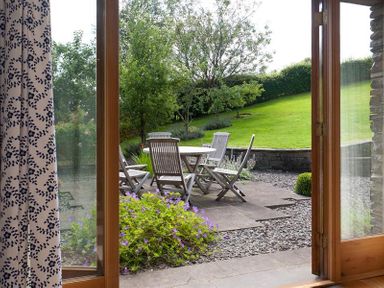 A garden patio with wooden chairs and a round table surrounded by plants and green grass at The Old Coach House in Troutbeck