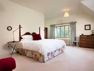 A bedroom with a double bed, bedside table with lamp, chest of drawers, chair, and window with curtains at The Old Coach House in Troutbeck