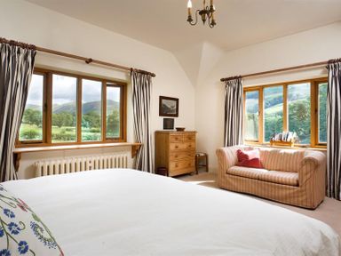 A bedroom with a bed a wooden dresser a striped sofa and windows with views of greenery at The Old Coach House in Troutbeck