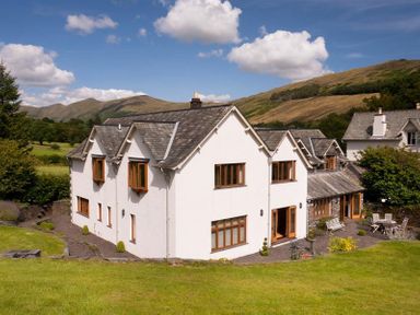 A white house with multiple windows and slate roof on a green lawn with hills in the background at The Old Coach House in Troutbeck