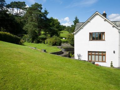A white house with brown windows next to a green lawn and trees at The Old Coach House in Troutbeck