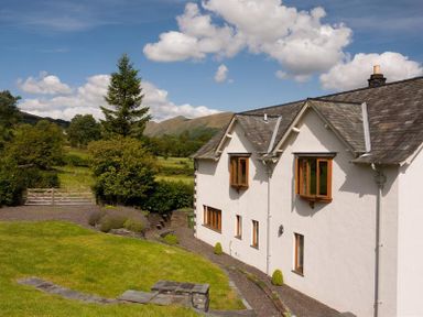A white house with brown windows surrounded by grass and trees with hills in the background at The Old Coach House in Troutbeck
