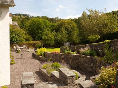 An outdoor garden with stone walls gravel paths a seating area with table and chairs and various plants and trees at The Old Coach House in Troutbeck