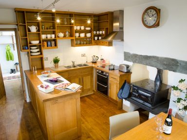 A kitchen with wooden cabinets and shelves with dishes a double sink an electric stove a toaster and a black stove with a stone beam above at Oakbank in Langdale