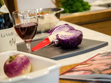 A kitchen countertop with a glass of red wine a bottle of wine a halved purple cabbage a turnip a cutting board with a knife and a cookbook at Oakbank in Langdale