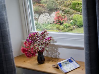 A window with curtains with flowers in a vase and a book on the windowsill overlooking a garden at Oakbank in Langdale