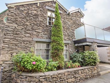 The exterior of a stone building with windows a small garden area with plants and flowers and a balcony with frosted glass panels at Workshop Cottage in Bowness
