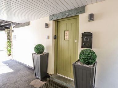 A green door with two grey planters with green plants and a black mailbox on a white wall at Workshop Cottage in Bowness