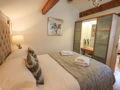A bedroom with a bed with pillows and folded towels a lamp on a bedside table a framed picture and sliding door wardrobe with a mirror and flowers at Workshop Cottage in Bowness