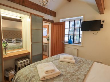 A bedroom with a bed, stacked towels, a window with blinds, a television mounted on the wall, a vanity with a mirror and chair at Workshop Cottage in Bowness