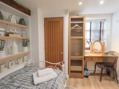 A bedroom with a bed towel folded on it wooden shelves with decor a wooden door a wardrobe and a wooden desk with a chair and mirror at Workshop Cottage in Bowness