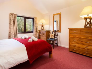 A bedroom with a bed covered by a red blanket and three rolled towels a wooden dresser a wooden chair with a cushion a wooden chest of drawers with a lamp and a mirror on the wall at Lakefield House in Hawkshead