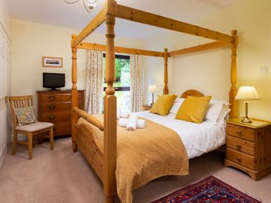 A bedroom with a wooden four-poster bed with yellow pillows and a blanket two bedside tables with lamps a wooden chest of drawers with a television a wooden chair and a window with curtains at Lakefield House in Hawkshead