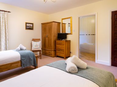 A bedroom with two single beds with towels on them a wooden wardrobe a chest of drawers with a TV a chair and a view into a bathroom at Lakefield House in Hawkshead