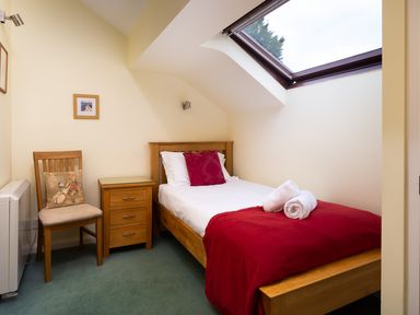 A small bedroom with a wooden single bed with red bedding and rolled white towels a wooden chair with a cushion and a wooden bedside table under a skylight window at Lakefield House in Hawkshead