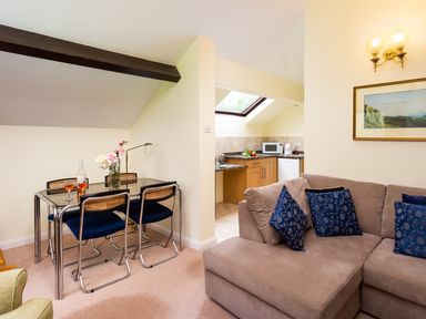 A dining area with a glass table and four chairs next to a beige sofa with blue cushions and a small kitchen with appliances at Lakefield House in Hawkshead