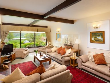 A living room with sofas, a coffee table, a model ship, framed paintings, and a large window showing a garden view at Lakefield House in Hawkshead