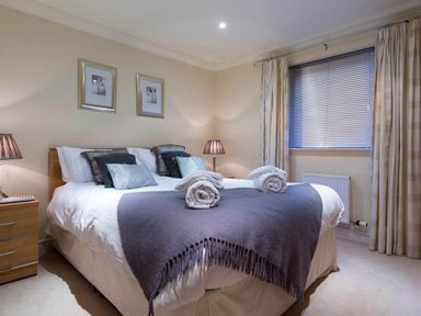 A bedroom with a bed with folded towels and cushions bedside tables with lamps and a window with blinds and curtains at Merewood Lodge in Ambleside