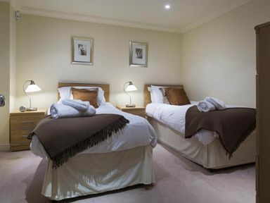 A bedroom with two single beds with brown blankets and pillows bedside tables with lamps and framed pictures on the wall at Merewood Lodge in Ambleside