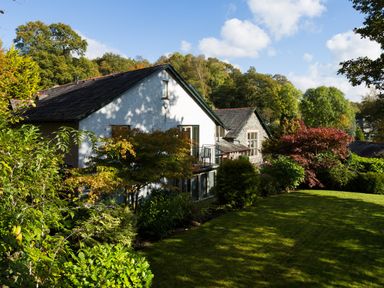 A house with white and stone walls surrounded by trees and a grassy lawn at Merewood Lodge in Ambleside
