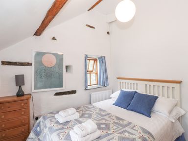 A bedroom with a bed, blue pillows, folded towels, a wooden chest of drawers, a lamp, a small window with curtains, and a wall picture at Cherry Tree Cottage at Satterthwaite in Hawkshead