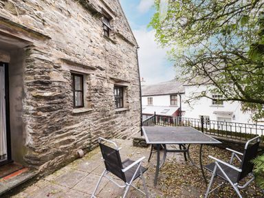 A stone patio area with a table and three chairs outside a building with stone walls and windows at Cherry Tree Cottage at Satterthwaite in Hawkshead
