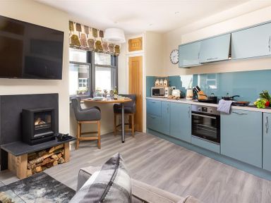 A kitchen with blue cabinets a dining table with chairs a wall-mounted TV and a fireplace at Old Wool Loft in Ambleside