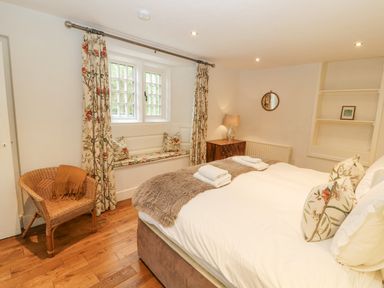 A bedroom with a bed, wooden chair, window seat, chest with lamp, and shelving at Home Farm House in Penrith