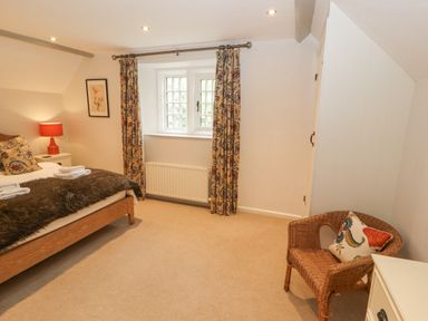 A bedroom with a bed, bedside table with lamp, window with curtains, wicker chair with cushion and a white dresser at Home Farm House in Penrith