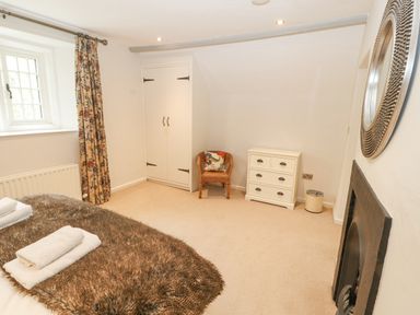 A bedroom with a bed covered by a fur blanket and towels a wicker chair a white chest of drawers a mirrored wall decor and a window with patterned curtains at Home Farm House in Penrith