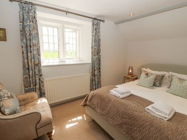 A bedroom with a double bed a chair beside a window with curtains and a bedside table with a lamp at Home Farm House in Penrith