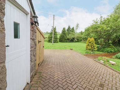 A paved driveway next to a stone wall with a white door and wooden gate leading to a grassy area with trees and shrubs at Home Farm House in Penrith