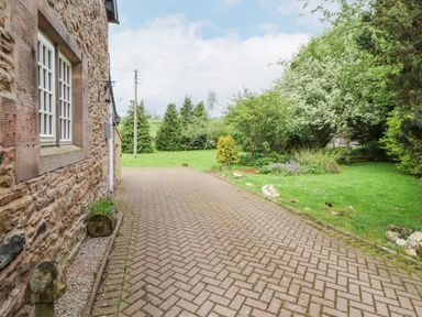 A stone exterior wall with windows next to a paved driveway and a garden with trees and grass at Home Farm House in Penrith