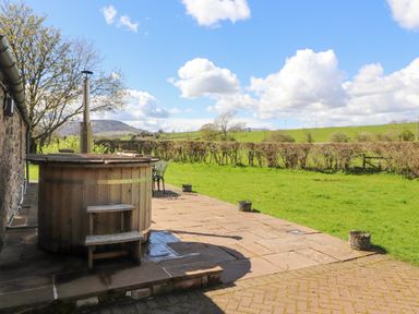 An outdoor area with a wooden hot tub on a stone patio next to a grassy lawn with fields and hills in the background at Home Farm House in Penrith