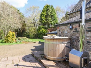 An outdoor wooden hot tub next to a stone building with a metal stove and chimney at Home Farm House in Penrith