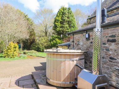 A wooden hot tub with a metal stove next to a stone building surrounded by trees at Home Farm House in Penrith