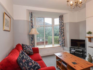 A living room with a red sofa blue floral curtains a wooden coffee table and a television at Fernleigh Cottage Ambleside