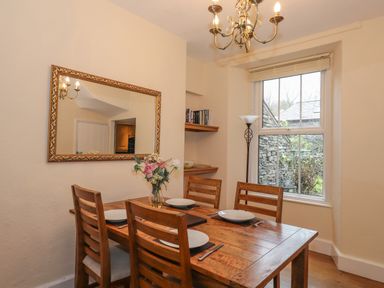 A dining room with a wooden table set for four people with plates and cutlery a flower vase on the table a large wall mirror a window and a floor lamp at Fernleigh Cottage in Ambleside