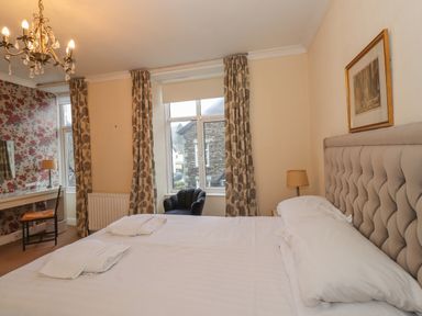 A bedroom with a large bed with white linens two towels on it a chandelier a desk with a chair floral curtains two windows and a framed picture on the wall at Fernleigh Cottage Ambleside