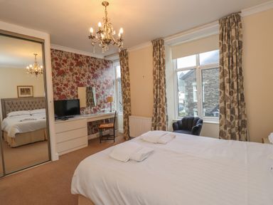 A bedroom with a bed, a dresser with a TV, a chair, floral curtains, and a window showing an outside building at Fernleigh Cottage in Ambleside