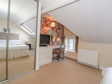 A bedroom with a bed reflected in a mirrored wardrobe, a desk with a television and chair, floral wallpaper, and a window at Fernleigh Cottage in Ambleside