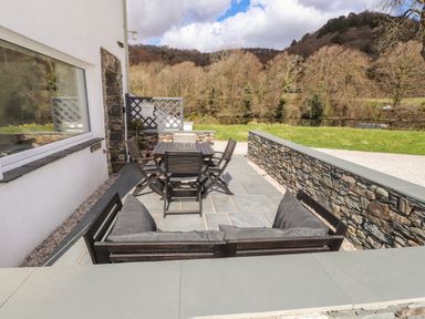 An outdoor patio with a wooden table, chairs, and a cushioned bench with a grass lawn and trees in the background at Riverside Hideaway in Newby Bridge