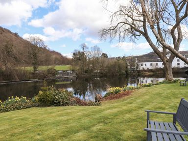 A garden with a tree and benches overlooking a river with buildings and trees in the background at Riverside Hideaway in Newby Bridge