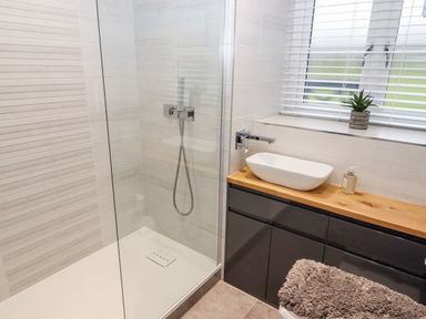A bathroom with a walk-in shower a wooden countertop with a white basin sink a soap dispenser a small plant on the windowsill and a gray cabinet below at Riverside Hideaway in Newby Bridge