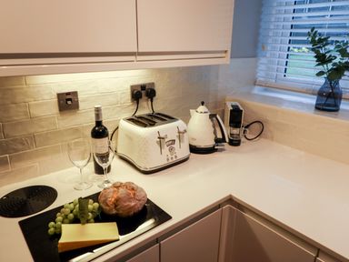 A kitchen countertop with a toaster, electric kettle, coffee machine, wine bottle, two glasses, bread, cheese, and grapes at Riverside Hideaway in Newby Bridge